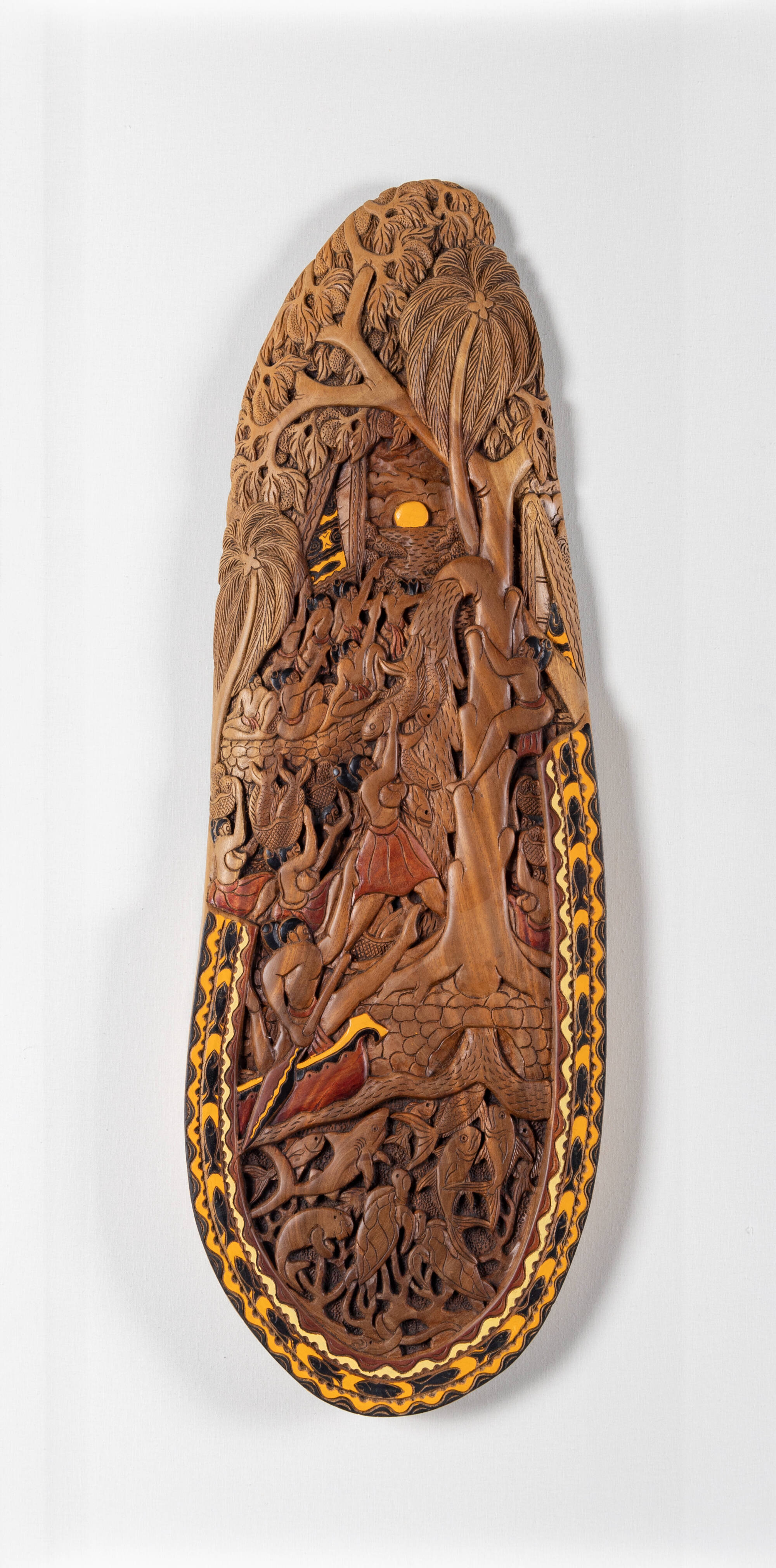 A piece of wood carved to depict many natives gathered around a tree which is spewing fish out of limb.